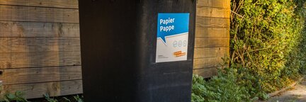 Schwarze Mülltonne mit blauem Deckel und Aufkleber 'Papier Pappe' steht auf Gehweg vor Holzzaun und Grünpflanzen | © Die Bremer Stadtreinigung