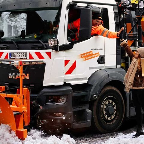 Schneeräumfahrzeug der Bremer Stadtreinigung mit Schneepflug auf verschneiter Straße neben einer Personen mit Schneeschieber, der Fahrer und die Person klatschen ab | © Die Bremer Stadtreinigung